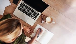 Top view shot of woman sitting at table with laptop and coffee writing on notebook updating financial information.