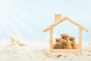 Vacation home estate planning. Wooden home model and money bag put on the beautiful sand and starfish on blue sky background in summer vacation, Saving or loan for buy a new house or real estate owner concept.