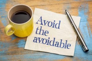 Avoid the Avoidable handwritten in ink on a napkin, next to a yellow coffee mug, on a distressed table.