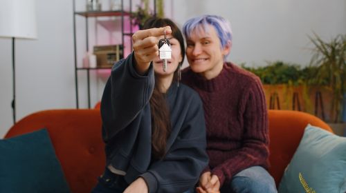 Two women sitting next to each other hugging while one holds up the keys to their new house.