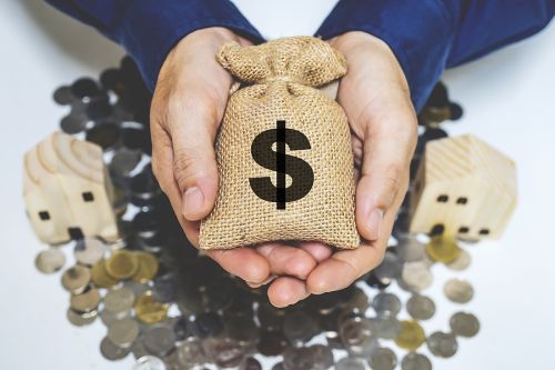 hands holding a brown burlap sack of money with coins and wooden houses in the background